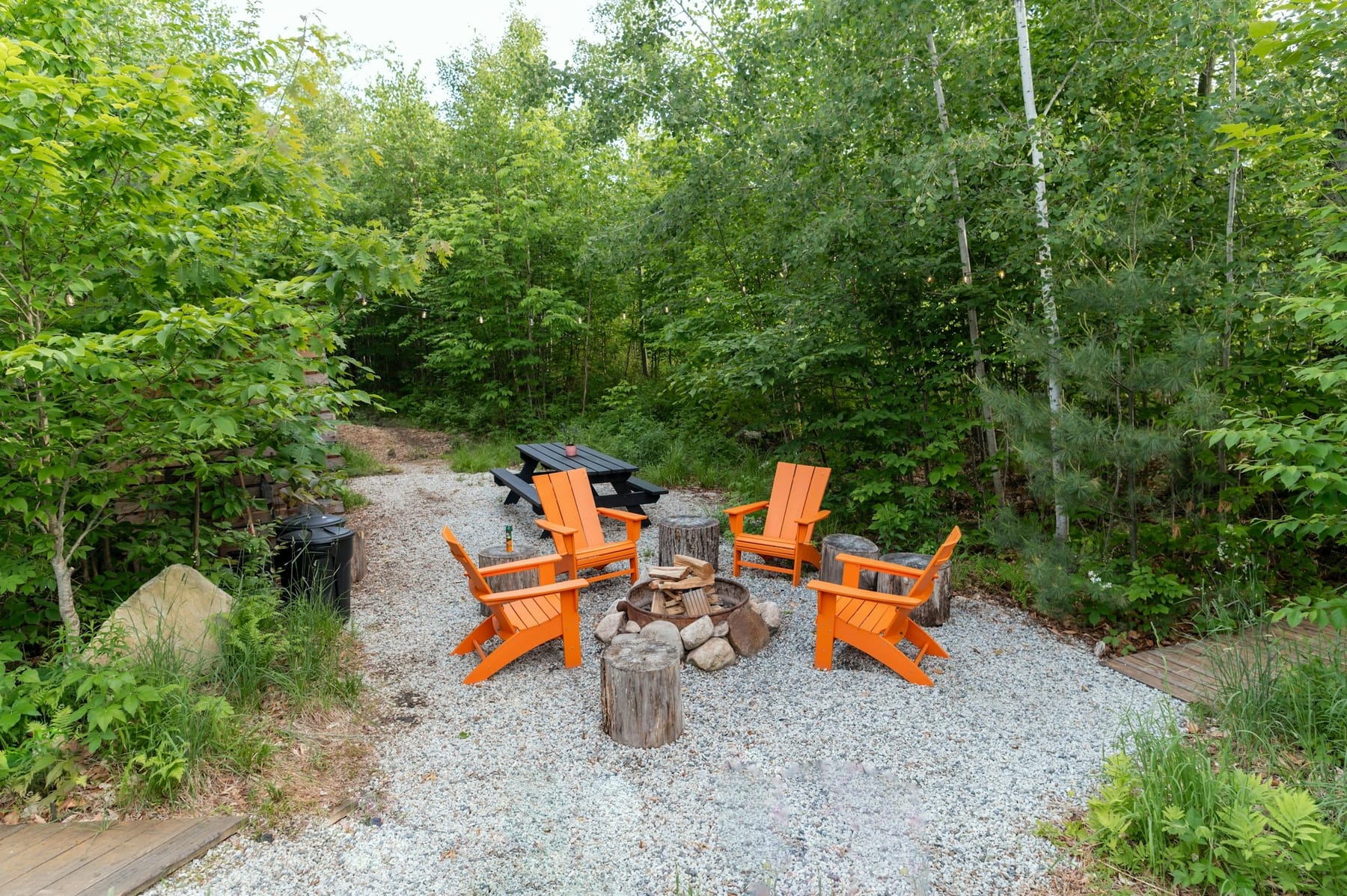 Adirondack chairs and a picnic table drawn up around a fire ring on a gravel clearing, with the tree line close on both sides.