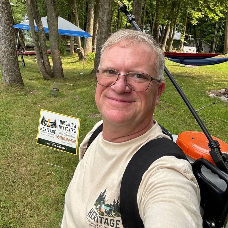 The owner of Heritage Outdoor Solutions in a company t-shirt with a backpack sprayer over one shoulder, standing in a shaded yard beside the firm's own sign; a hammock and a canopy are set up behind him.